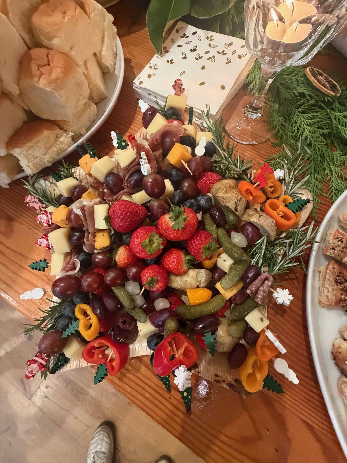 A holiday grazing board with strawberries, grapes, cheeses, cured meats, pickles, and rolls beside a wine glass