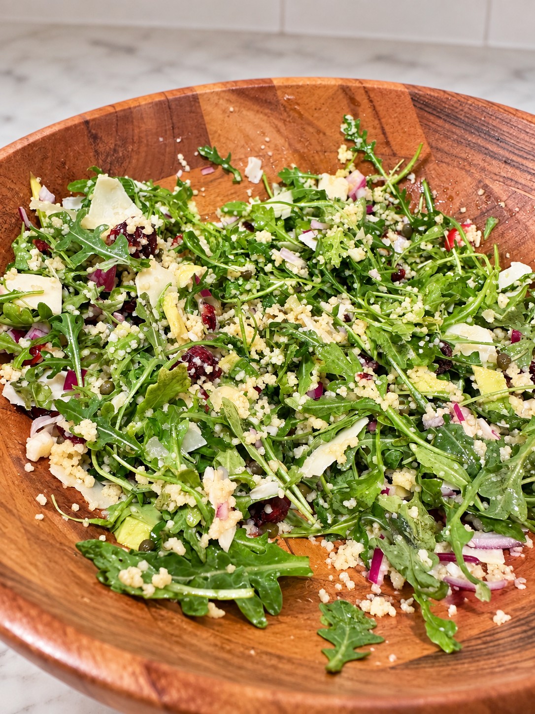 Arugula and regular couscous salad with dried cranberries, shaved Parmesan, and capers in a wooden bowl.