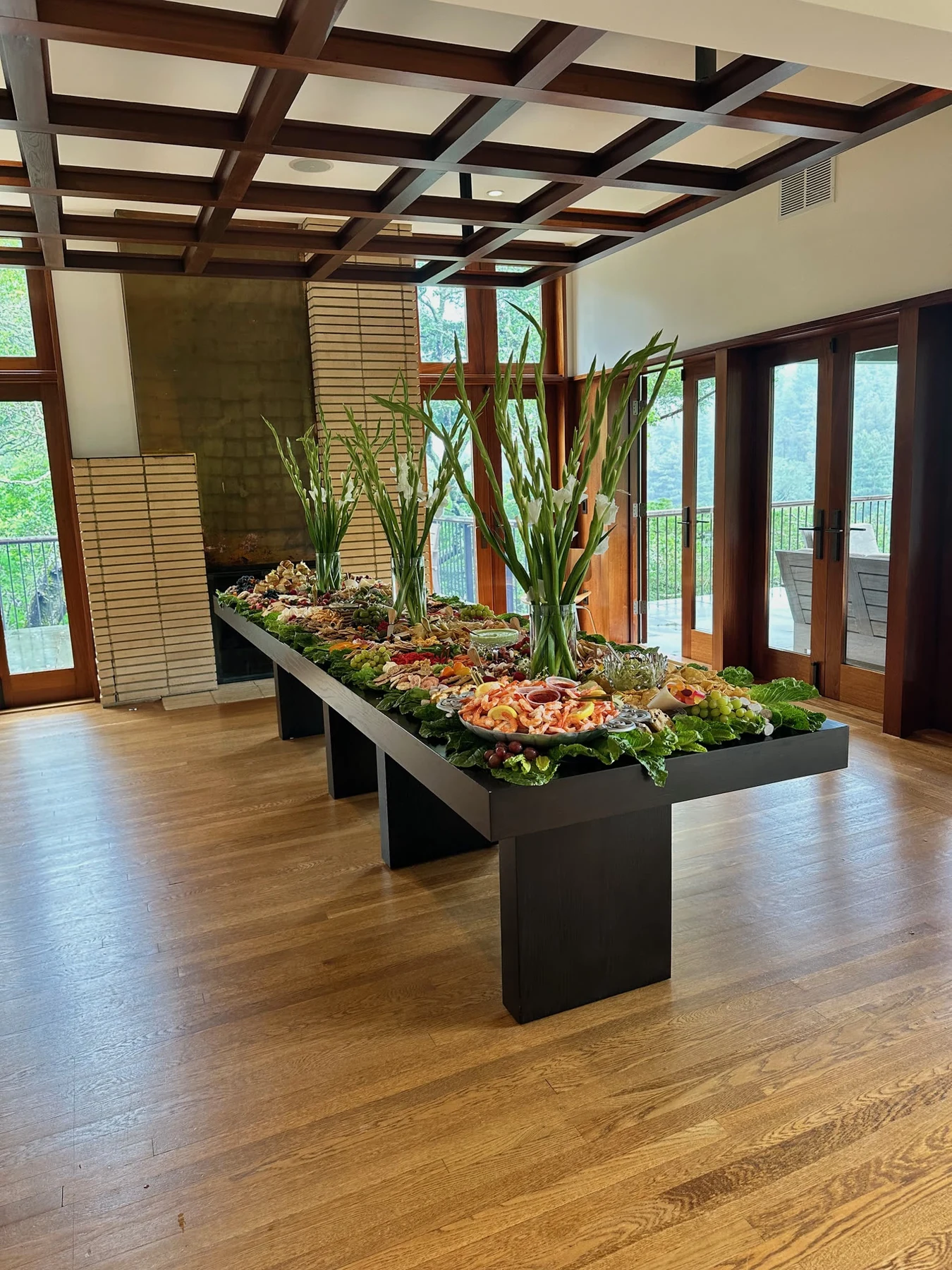 A pulled-back view of the complete black prom table with cascading leafy borders and tall white gladiolus in a timber-windowed room