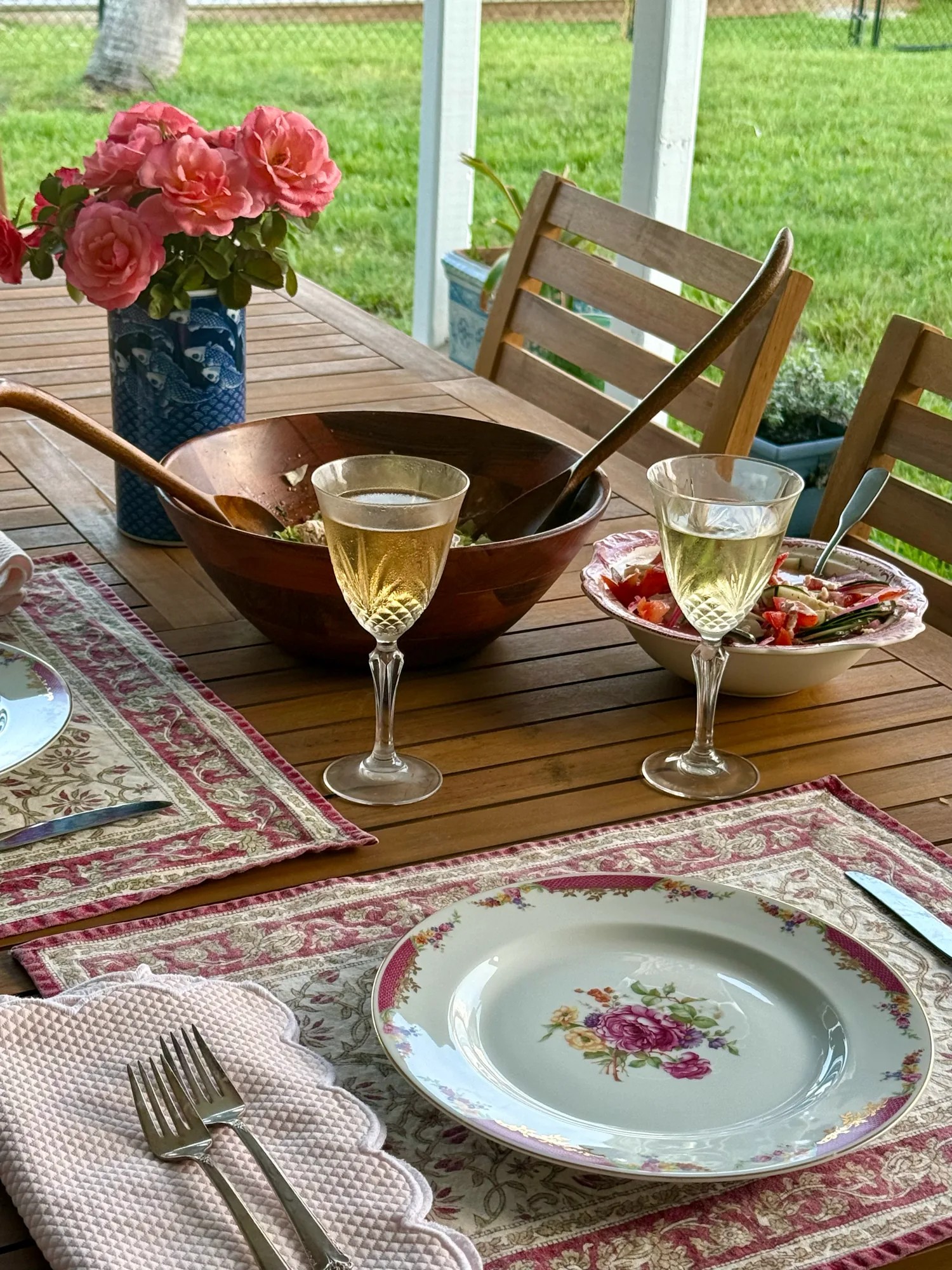 A garden place setting with floral china, patterned linens, wine glasses, salad bowls, and pink roses