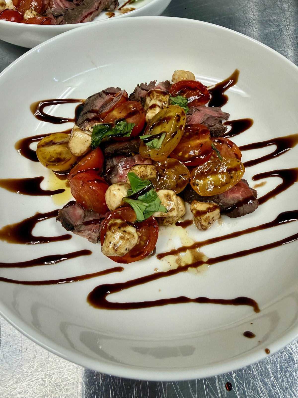 Sliced steak with roasted tomatoes and mozzarella, finished with basil and a balsamic drizzle on a white plate
