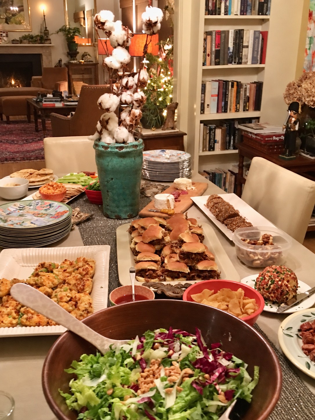 A holiday buffet with a turquoise vase of cotton stems, sliders, salad, appetizers, and stacks of patterned plates.