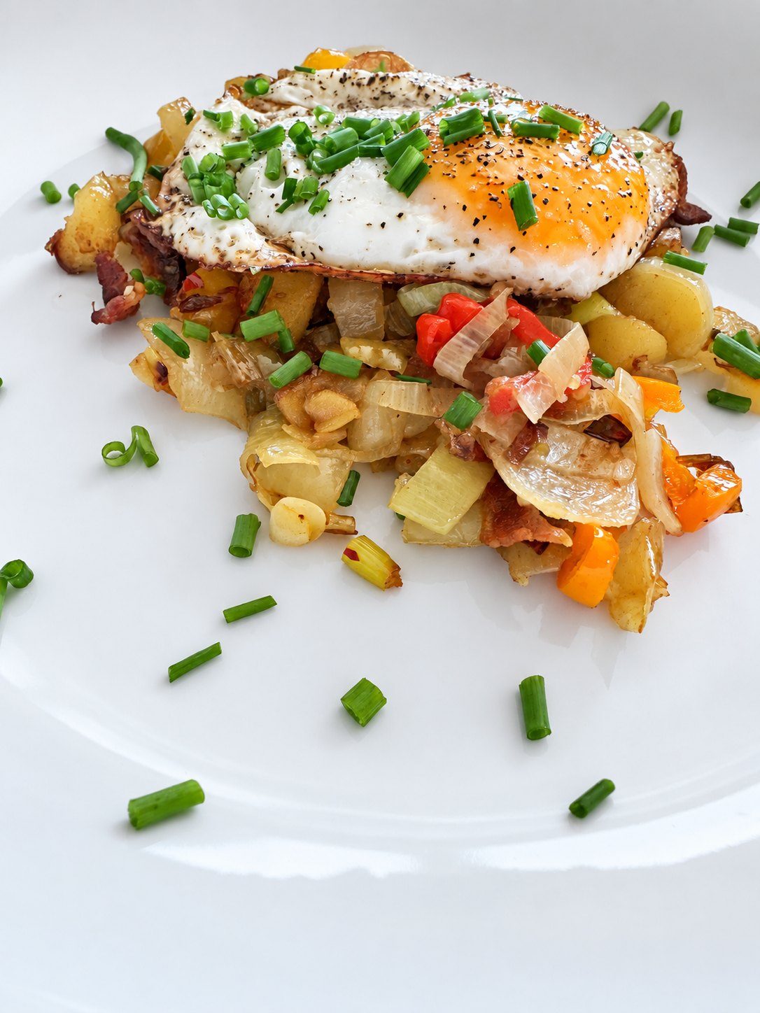 A fried egg with a bright yolk over a potato, onion, and pepper hash scattered with chopped chives on a white plate