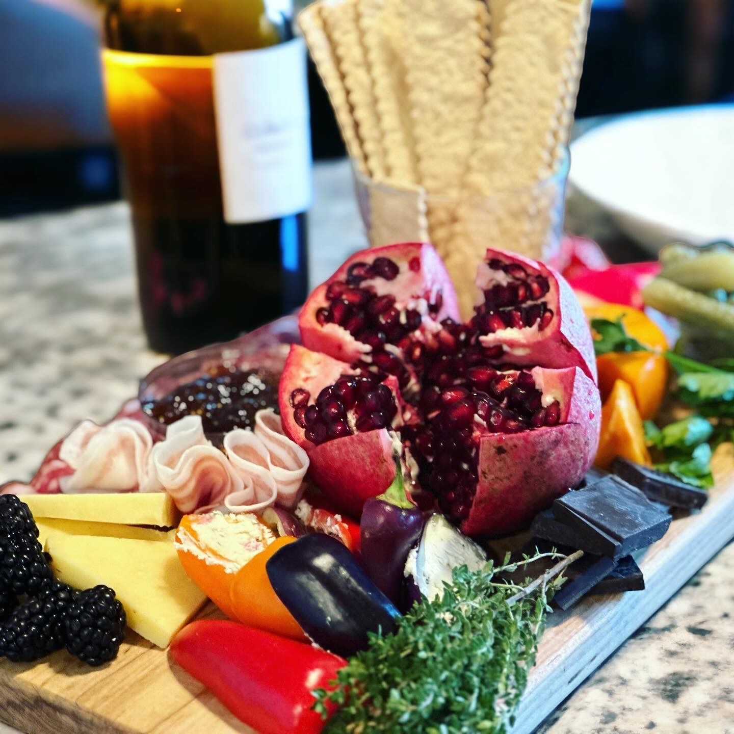 A cut pomegranate surrounded by cheese, blackberries, herbs, peppers, chocolate, and crackers