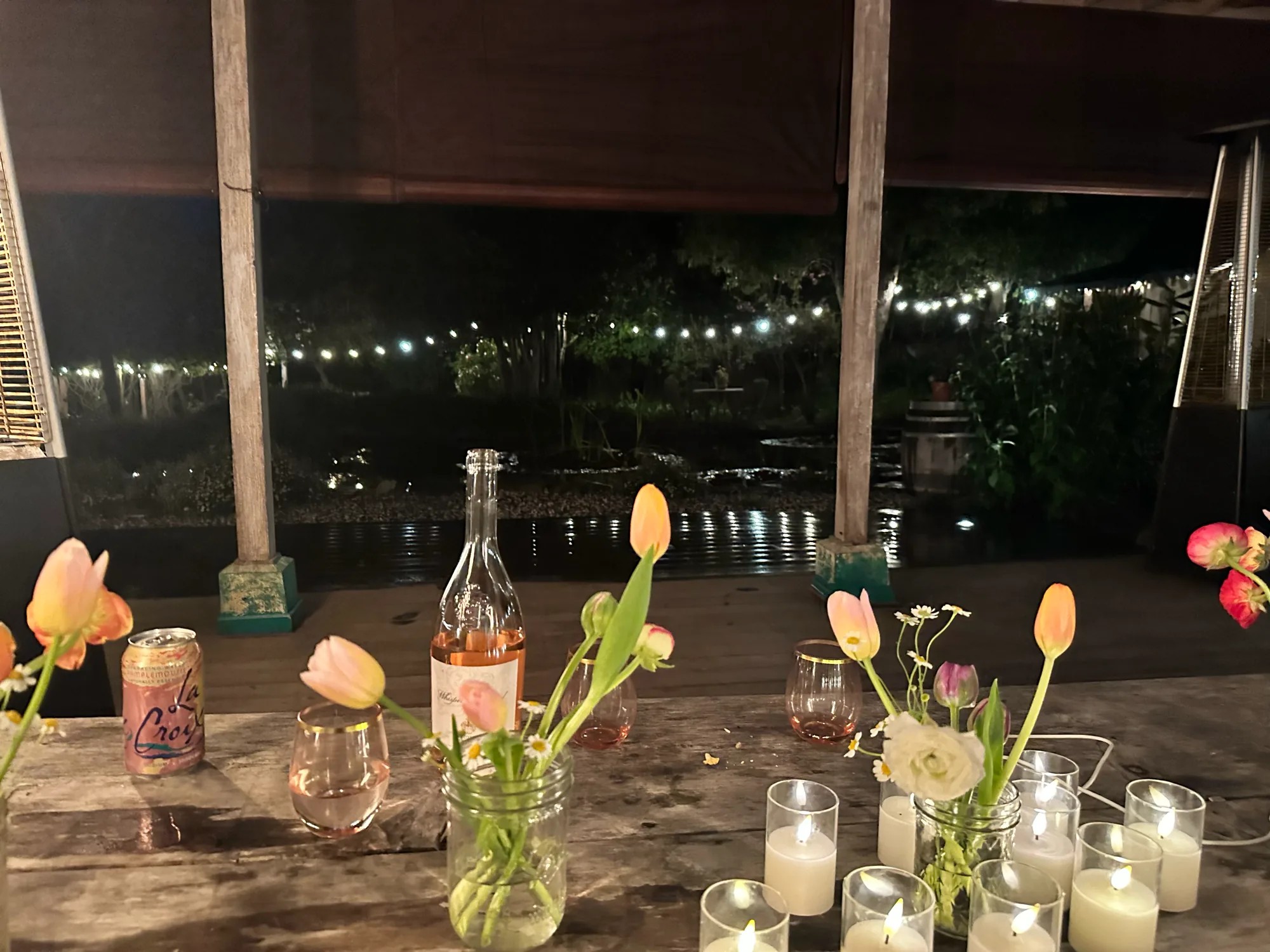 A weathered table glowing with candles, tulips, wine glasses, and garden string lights after dark