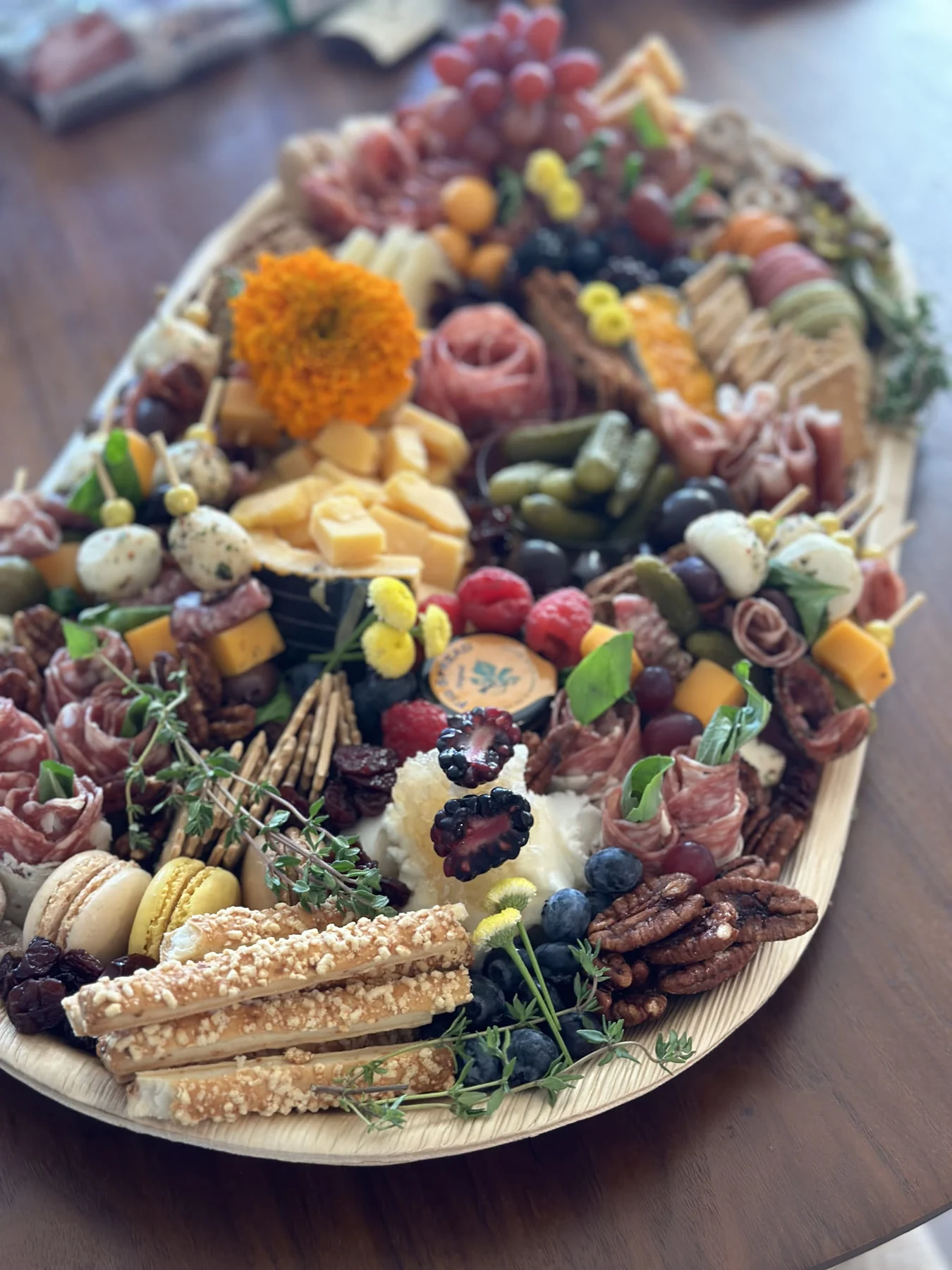 A large oval board layered with cheeses, cured meats, fruit, crackers, nuts, olives, and edible flowers