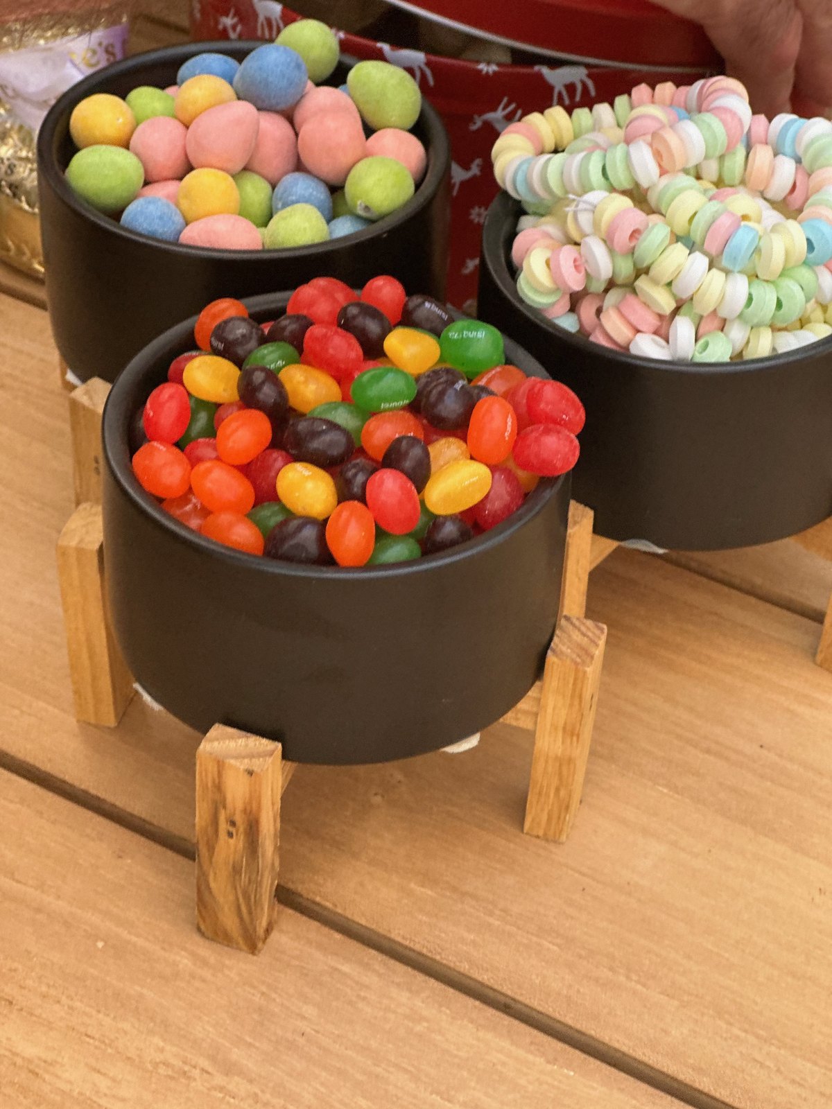 Three dark ceramic bowls on wooden stands filled with jelly beans, pastel candies, and candy necklaces