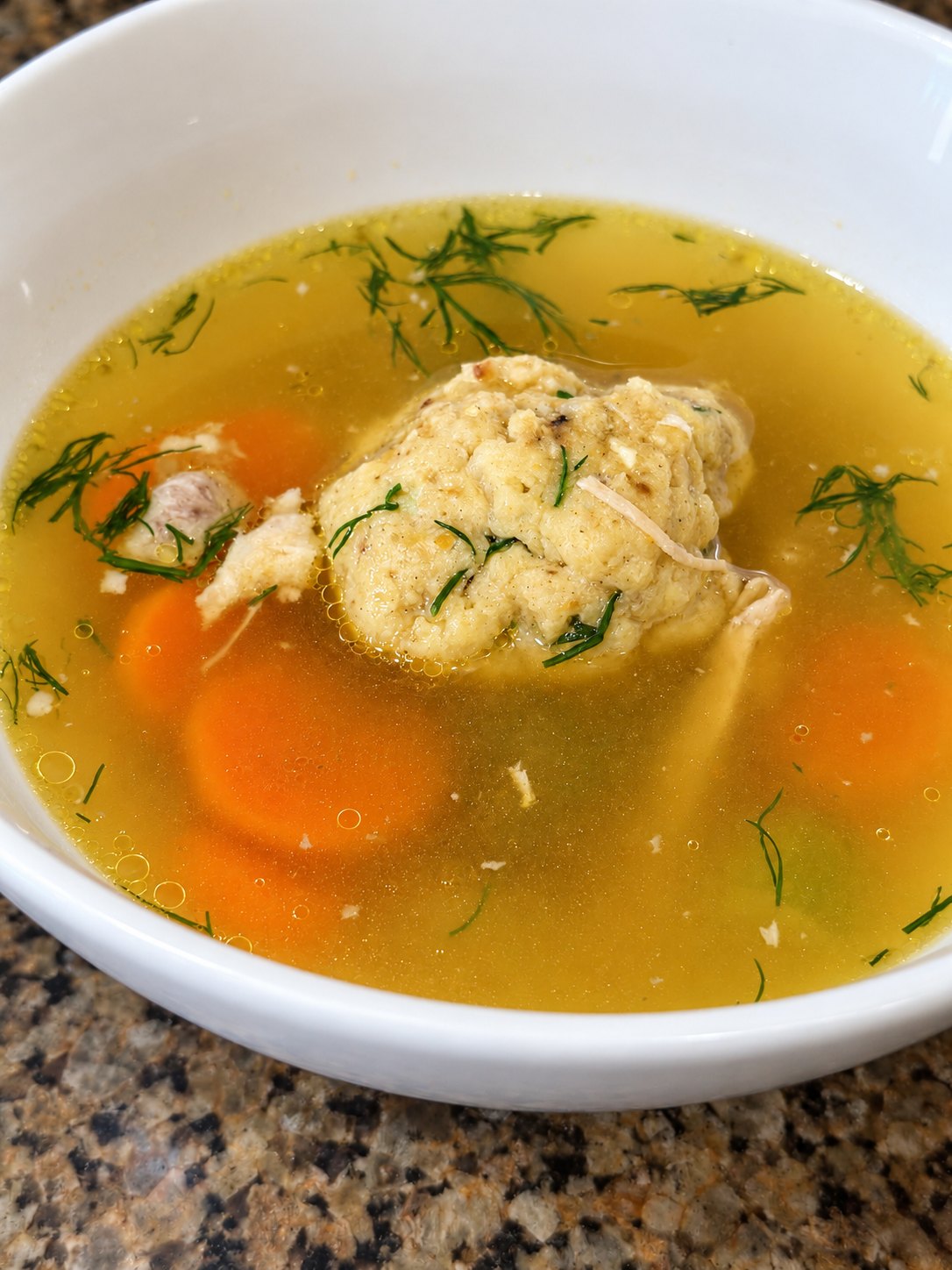 A white bowl of golden chicken broth with a matzo ball, sliced carrots, shredded chicken, and fresh dill
