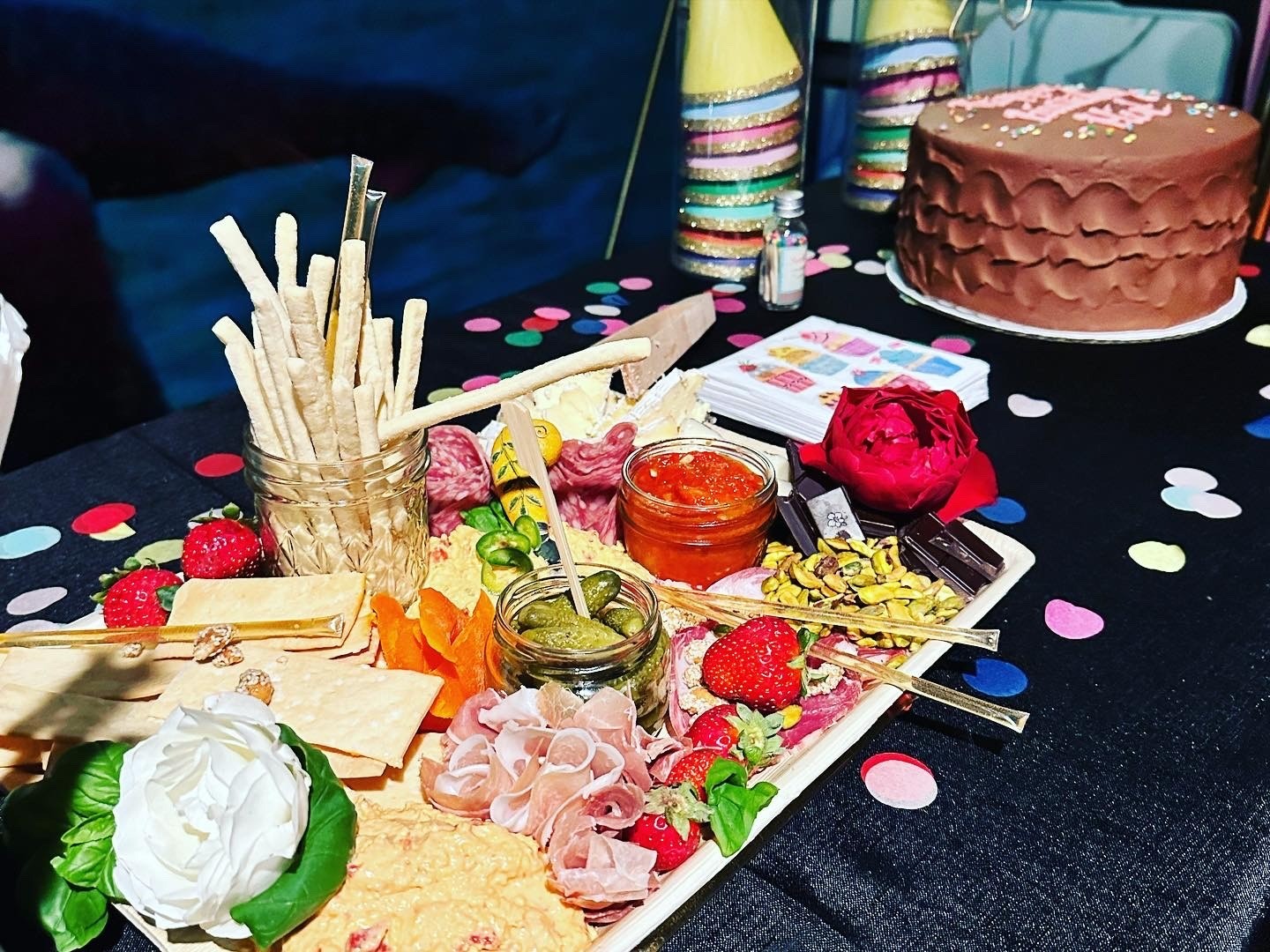 A colorful birthday spread with a grazing board, chocolate cake, roses, party hats, and confetti