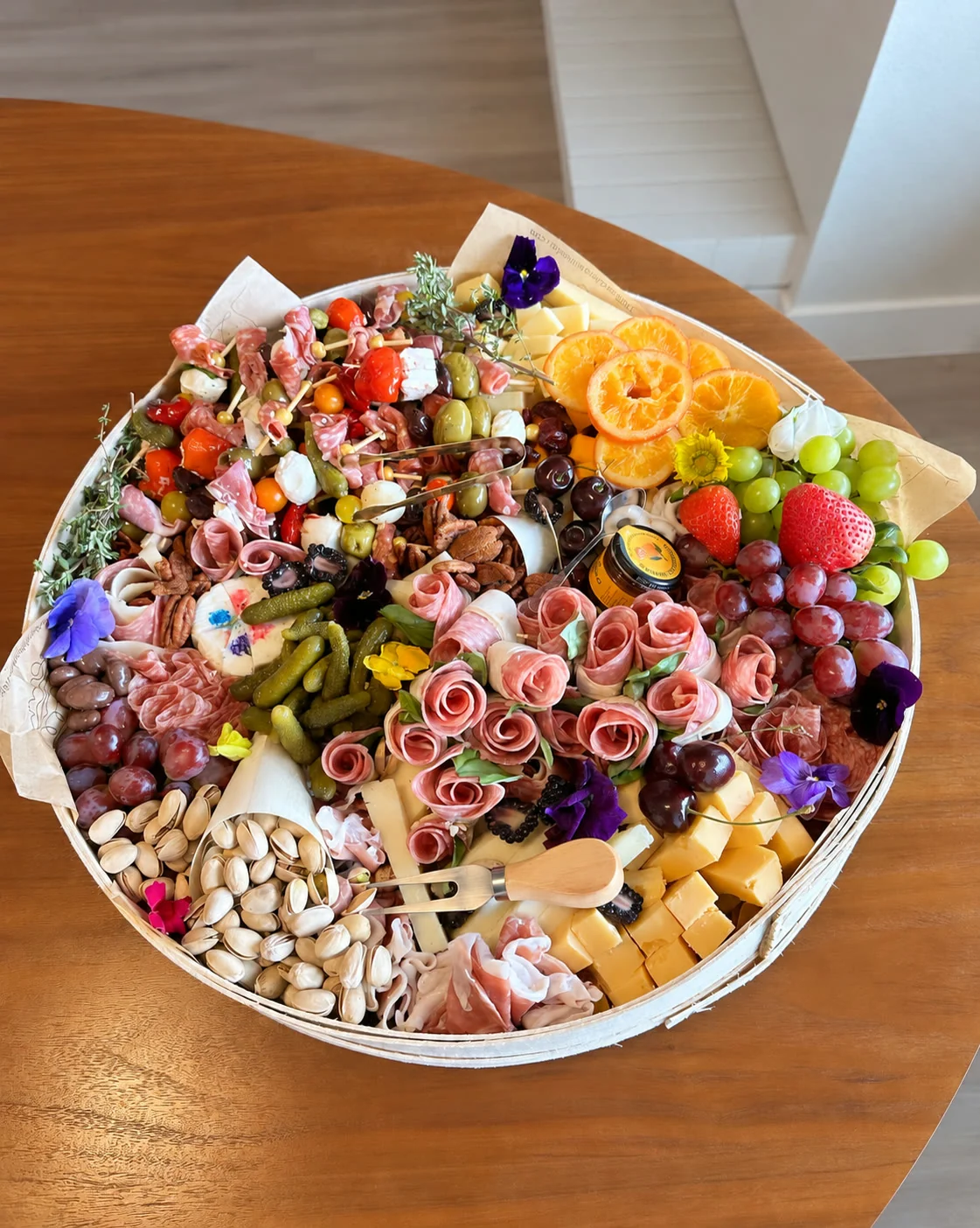 A round abundant charcuterie board with rolled cured meats, cheeses, fruit, nuts, crackers, and edible flowers