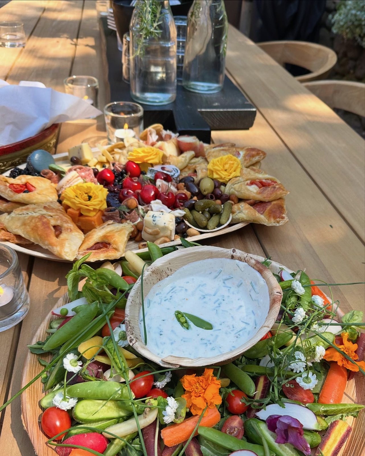 An outdoor table with a crudités and dip board and a pastry and cheese board dressed with fresh flowers