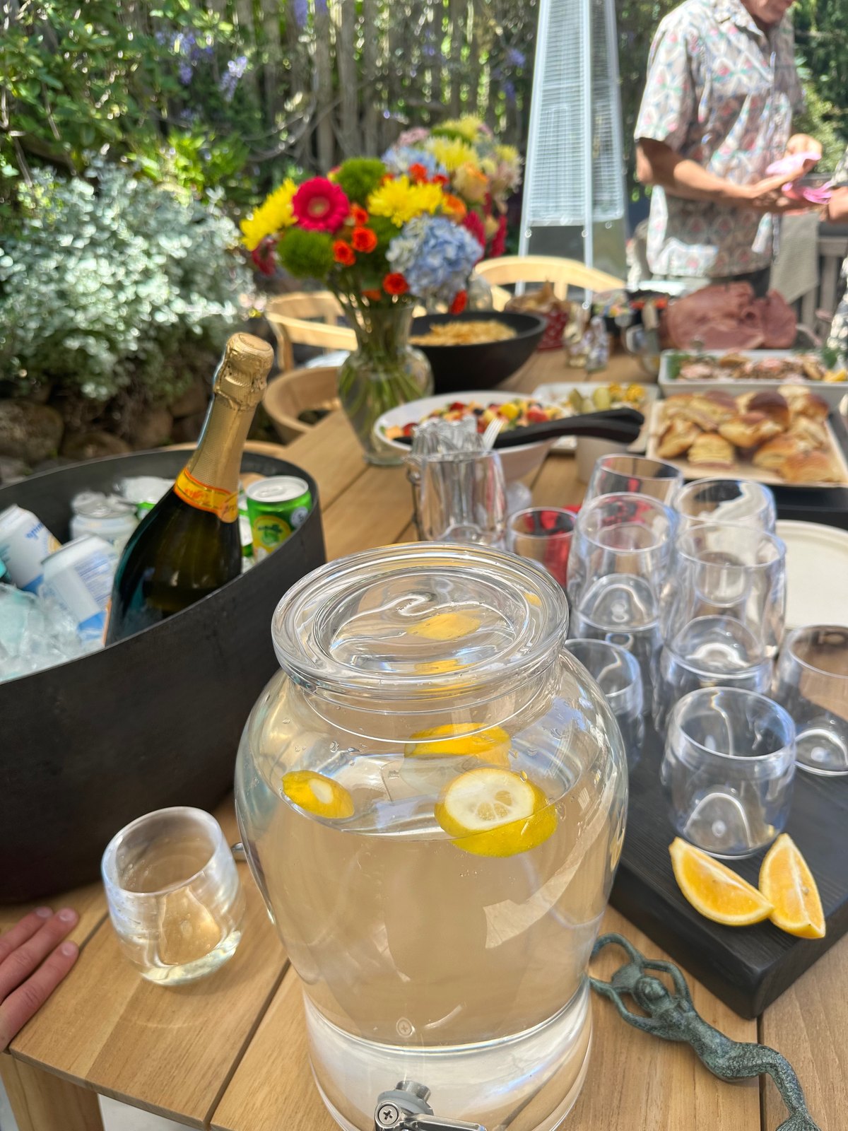 A glass dispenser of lemonade with lemon slices beside champagne on ice, glassware, and flowers on a garden buffet