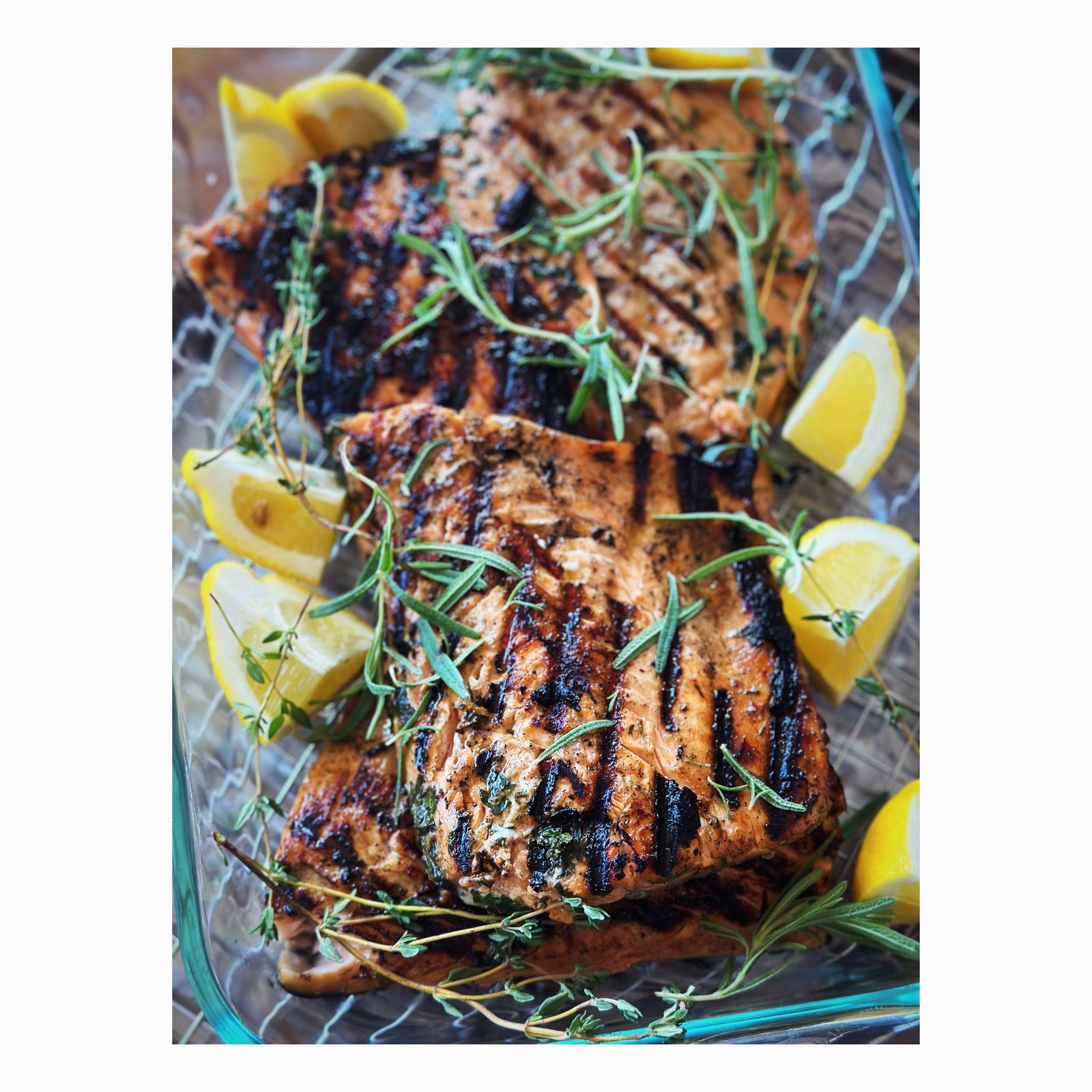 Grilled salmon fillets with char marks, topped with rosemary and thyme, lemon wedges alongside