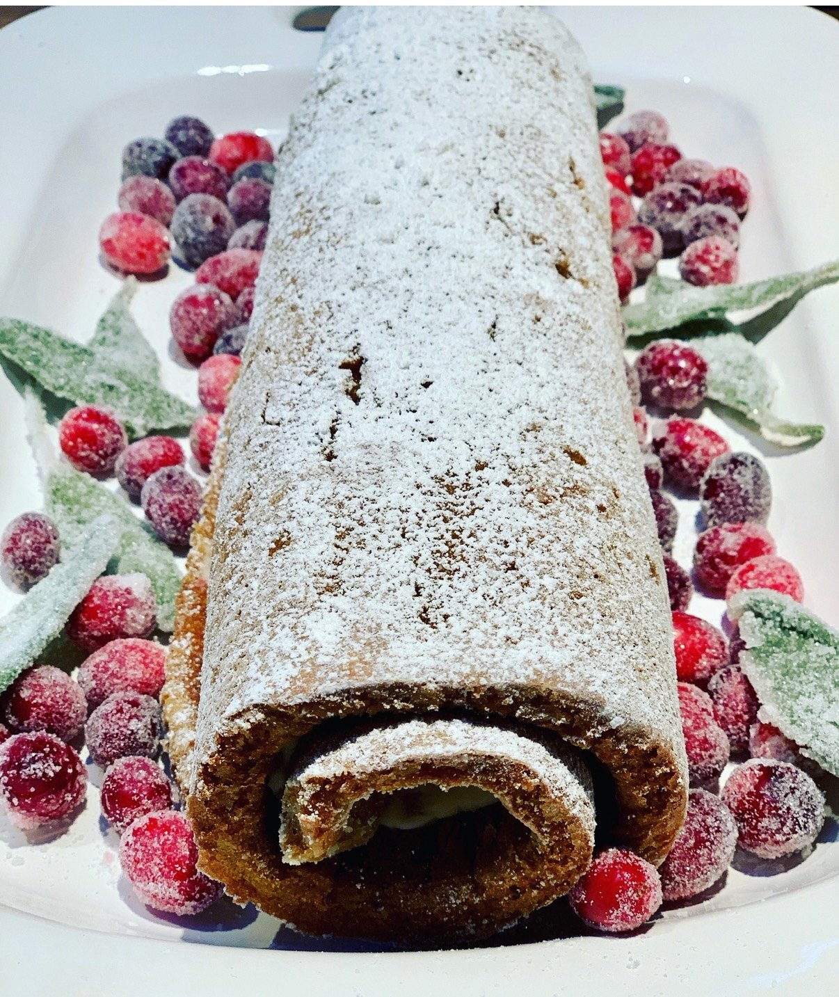 A powdered-sugar-dusted rolled cake surrounded by sugared cranberries and blueberries on a white platter