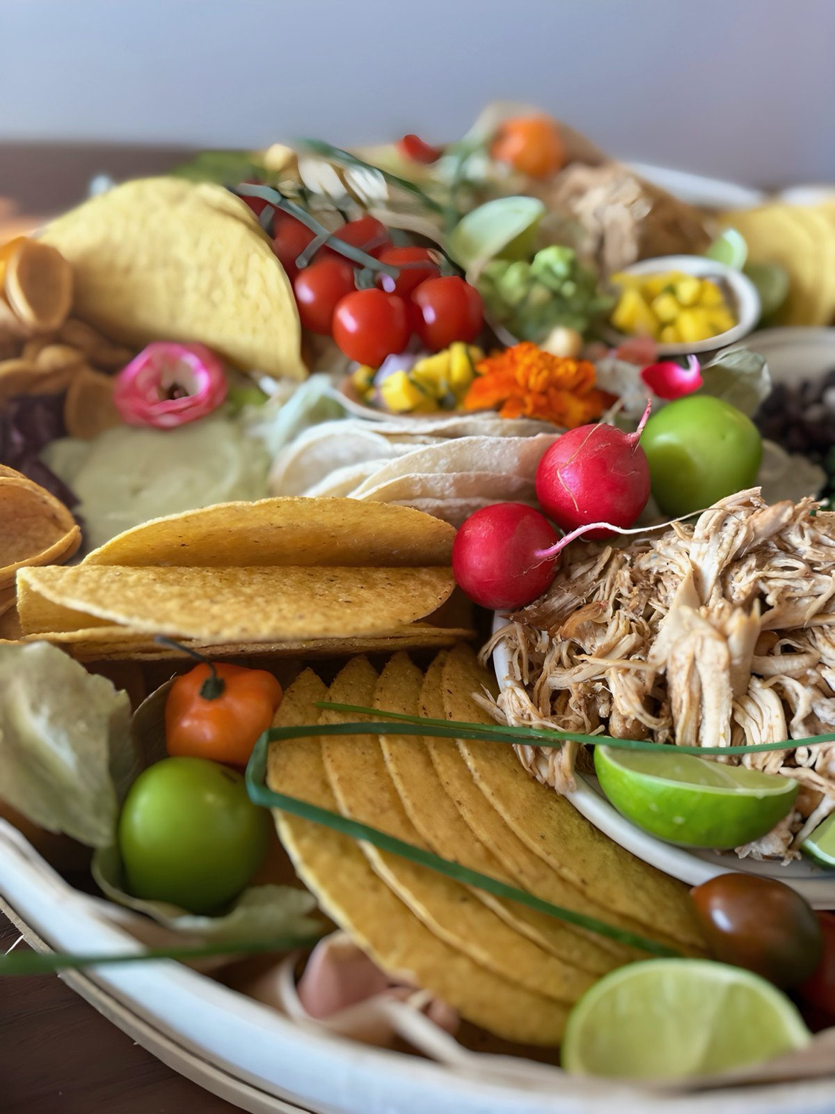 A taco board with crisp shells, shredded chicken, radishes, limes, cherry tomatoes, and edible flowers
