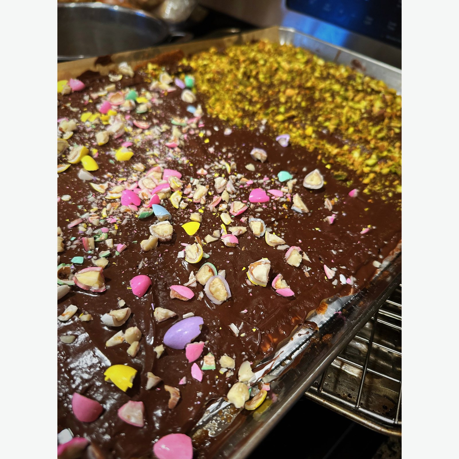 A sheet pan of dark chocolate bark scattered with chopped nuts, pistachios, and pastel candy shards