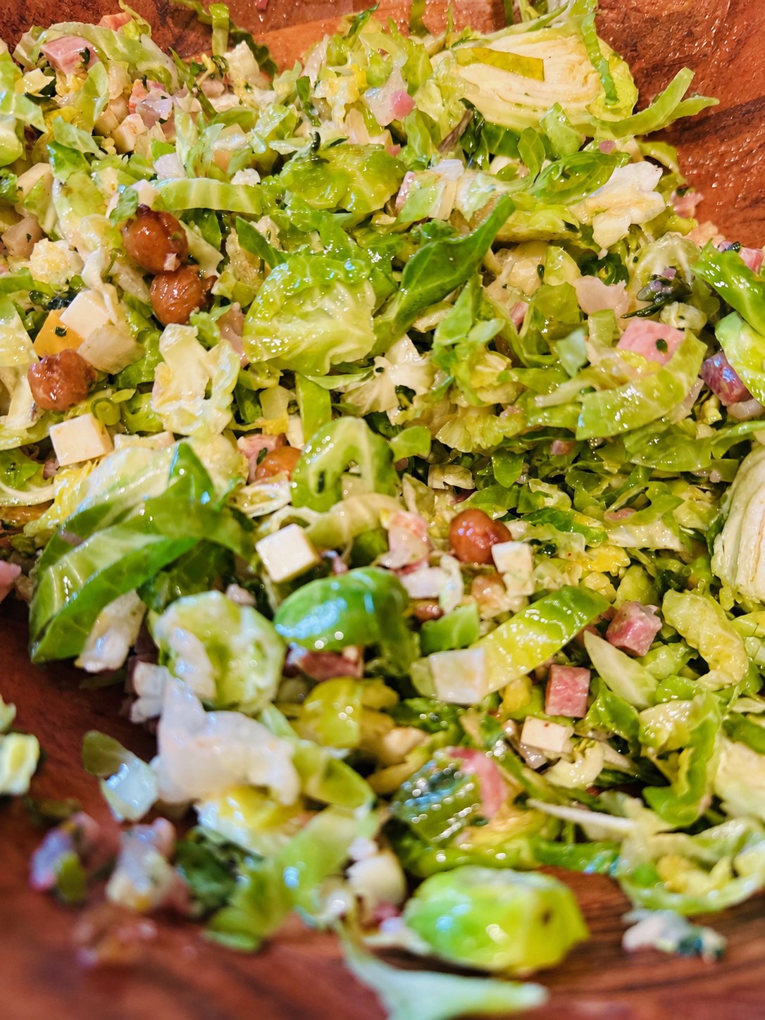 A finely chopped salad of shaved brussels sprouts and lettuces with diced salami, cheese, and glazed nuts in a wooden bowl