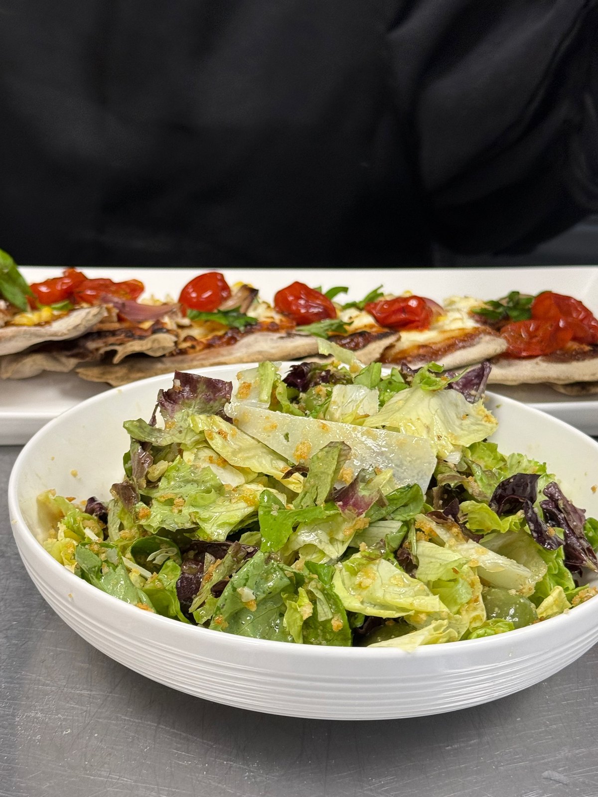 A bowl of mixed green salad in front of flatbreads topped with tomatoes and cheese
