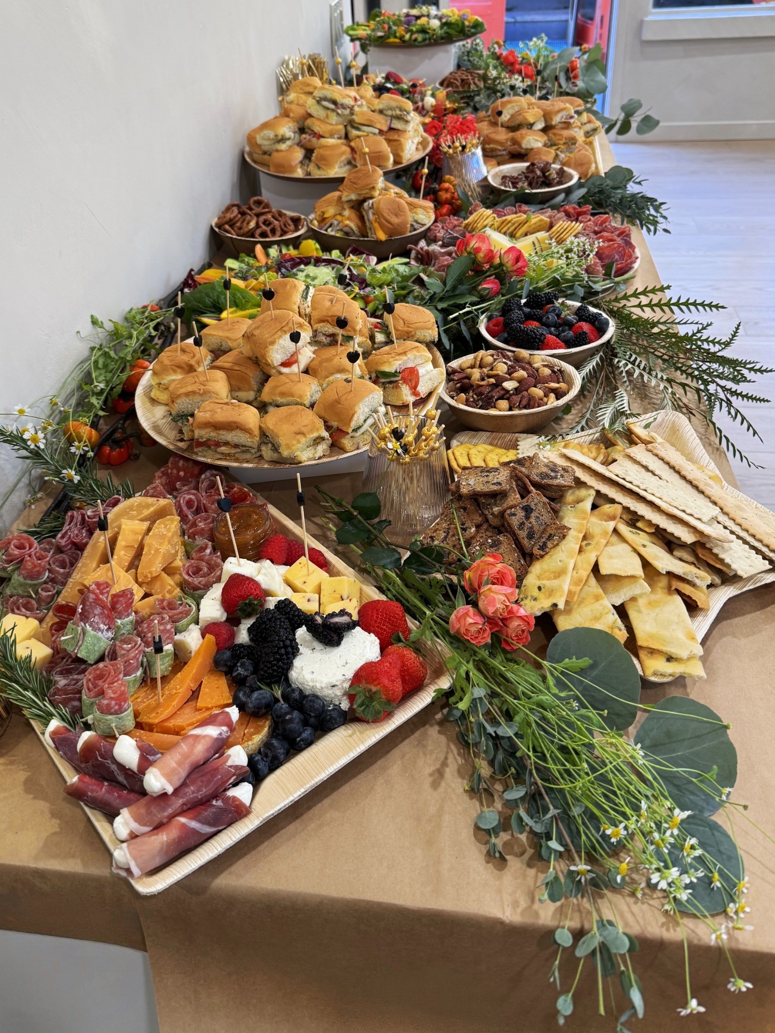 A long grazing table filled with sandwiches, cheeses, charcuterie, berries, crackers, nuts, and flowers