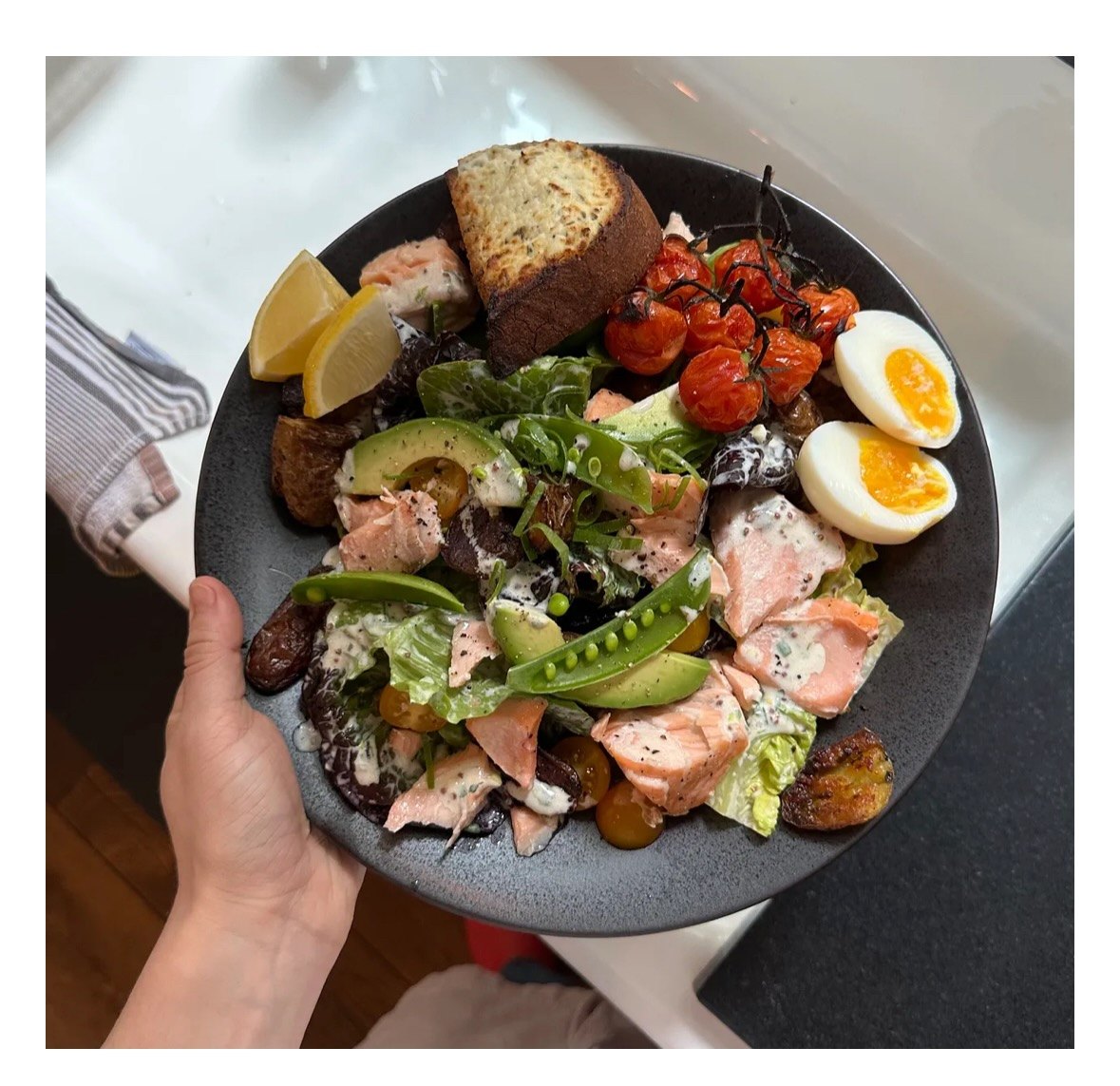 A hand holding a dark bowl of salmon, avocado, roasted tomatoes, jammy eggs, potatoes, and grilled bread