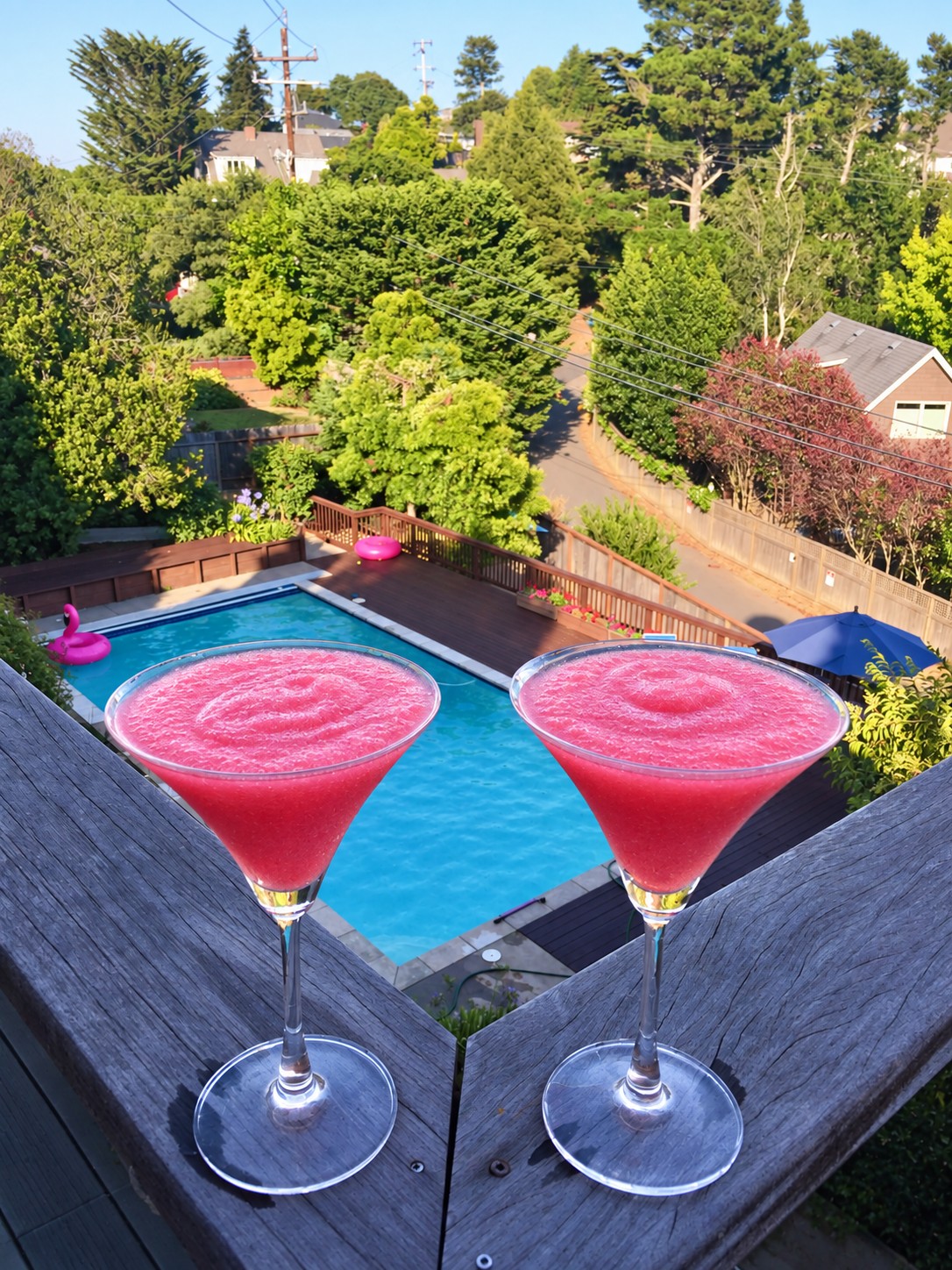 Two bright pink frozen drinks in stemmed glasses overlooking a swimming pool and green neighborhood.