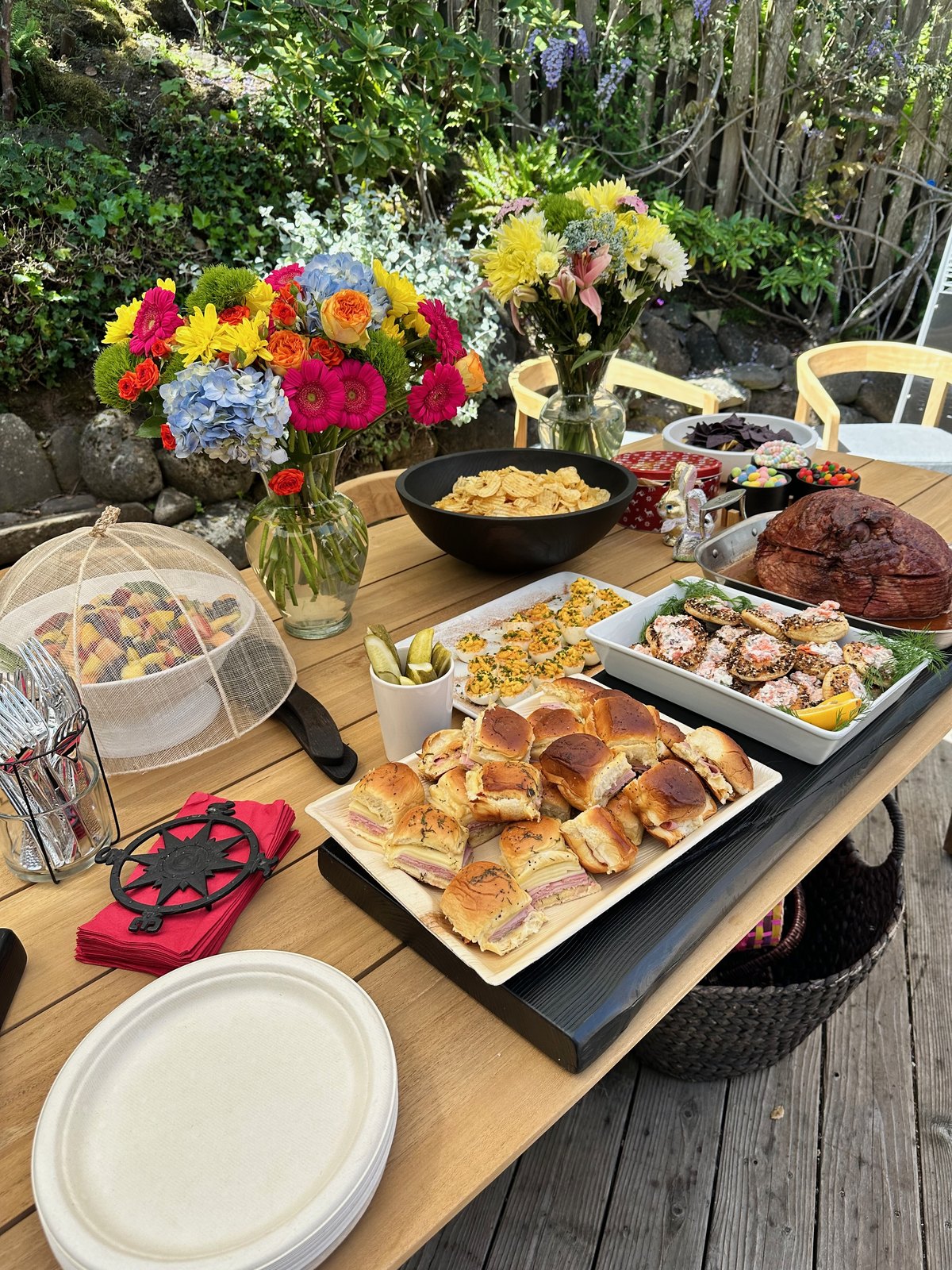 A wooden garden table laid with ham, sliders, deviled eggs, salmon bagel bites, fruit, and two bright bouquets