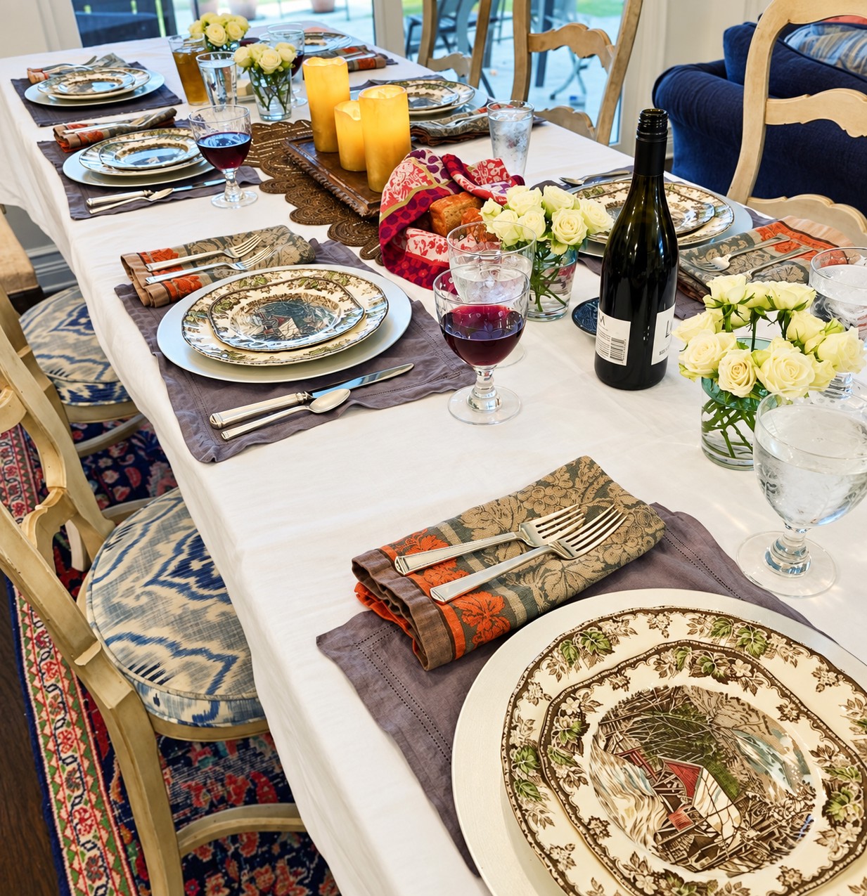 A dining table set with patterned plates, folded napkins, candles, wine, and small arrangements of white roses.