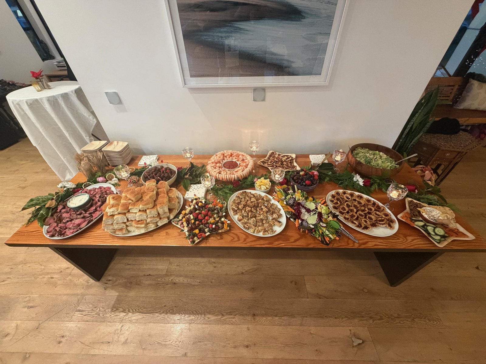 A long buffet table filled with platters of shrimp, sliced beef, rolls, salads, and small bites beneath a framed painting