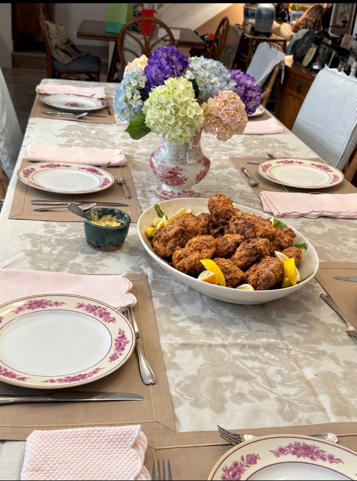 A vase of blue, purple, green, and blush hydrangeas on a set dinner table with a platter of fried chicken and lemon