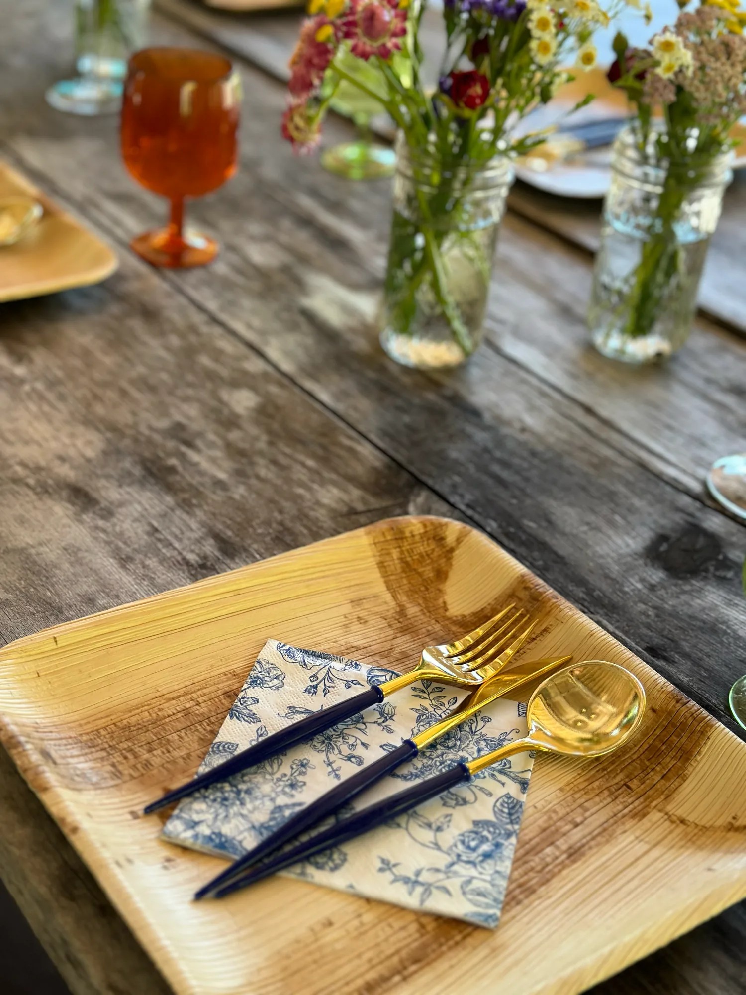 A palm-leaf plate set with a blue floral napkin and gold flatware on a weathered table with wildflowers