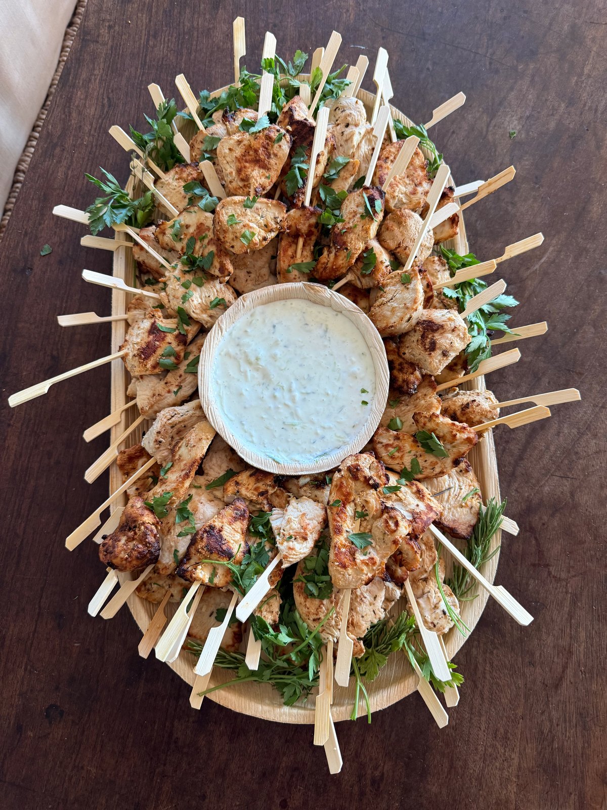 Grilled chicken skewers with parsley arranged around a bowl of herb dip on a round board