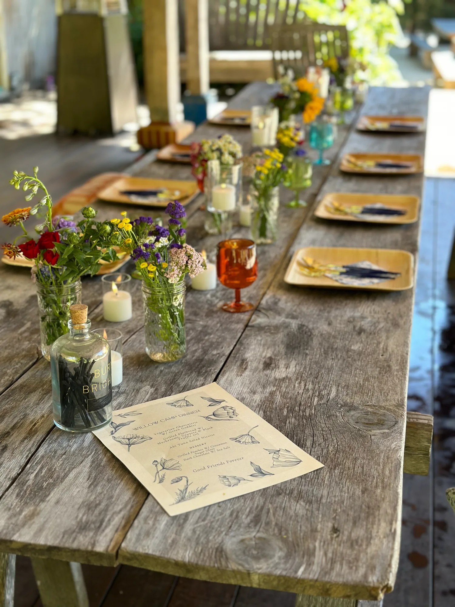 A long weathered garden table set with small jars of wildflowers, candles, plates, and a printed menu
