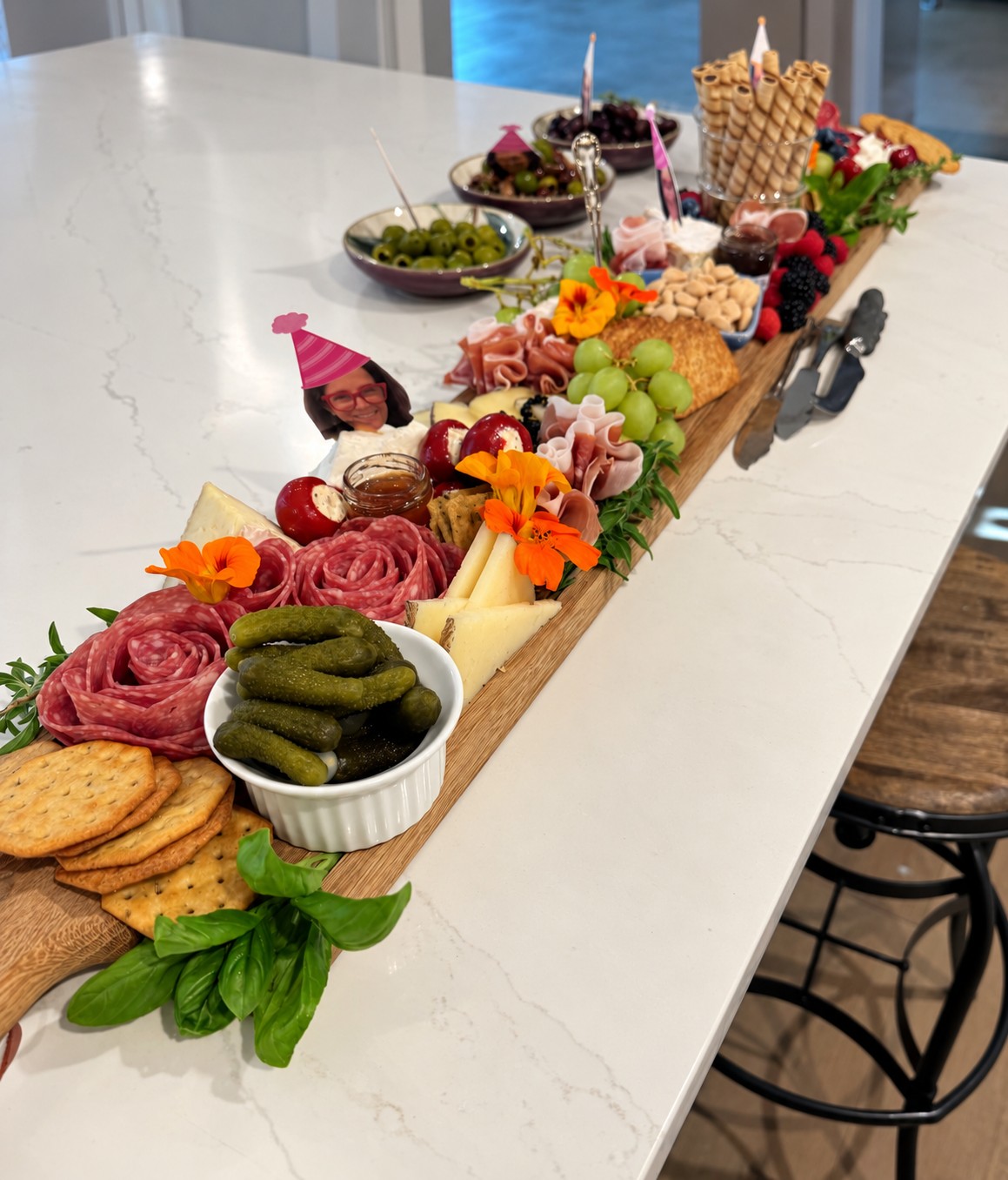 A long narrow party grazing board with salami roses, cheeses, grapes, berries, pickles, crackers, nuts, and edible flowers.