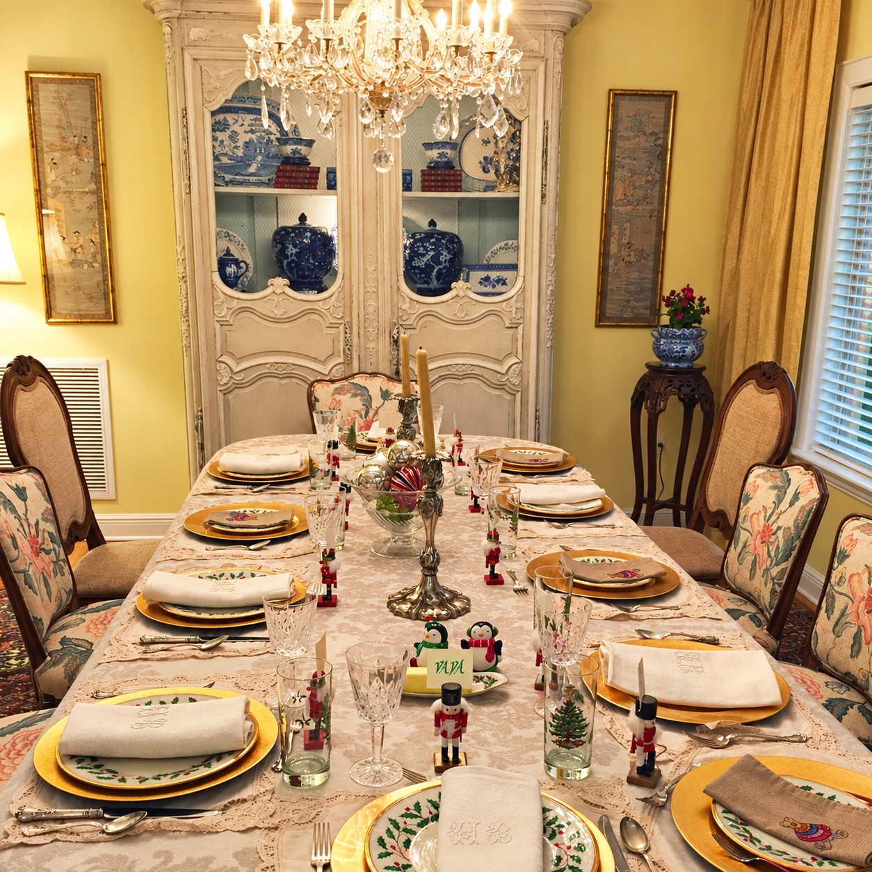 A holiday dining table set with gold chargers, patterned china, crystal glasses, candles, nutcrackers, and embroidered napkins beneath a chandelier.