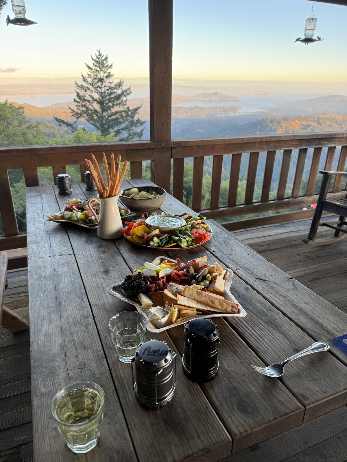 A weathered deck table set with charcuterie, crudités, and drinks overlooking mountains at sunset
