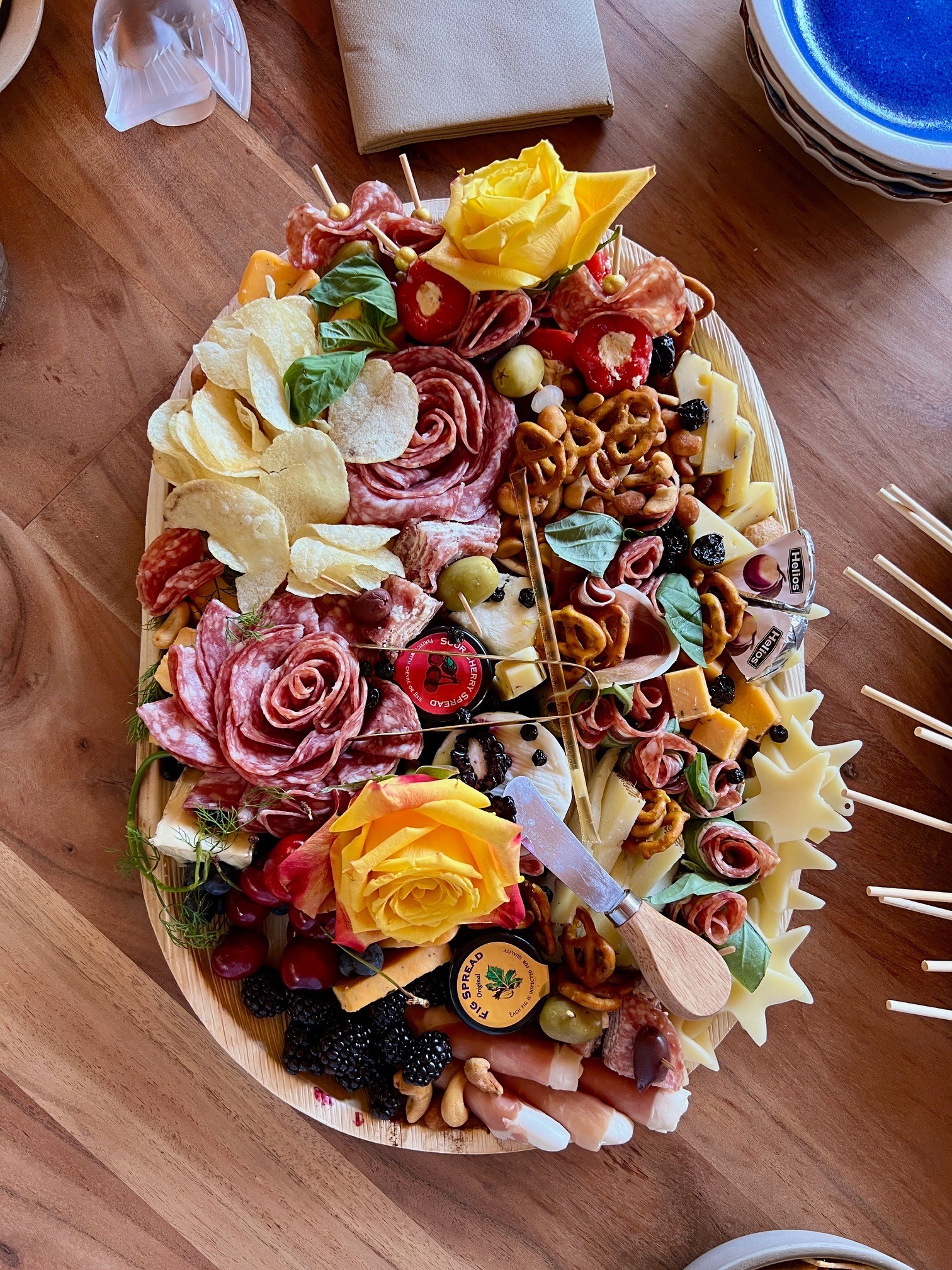 An abundant oval board with cheeses, cured meats shaped into roses, fruit, nuts, pretzels, and yellow roses