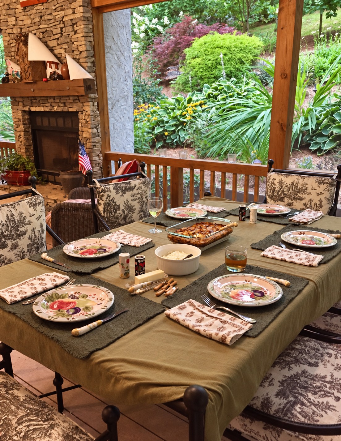 A dinner table set with floral plates beside a stone fireplace and a wide window overlooking a green garden.