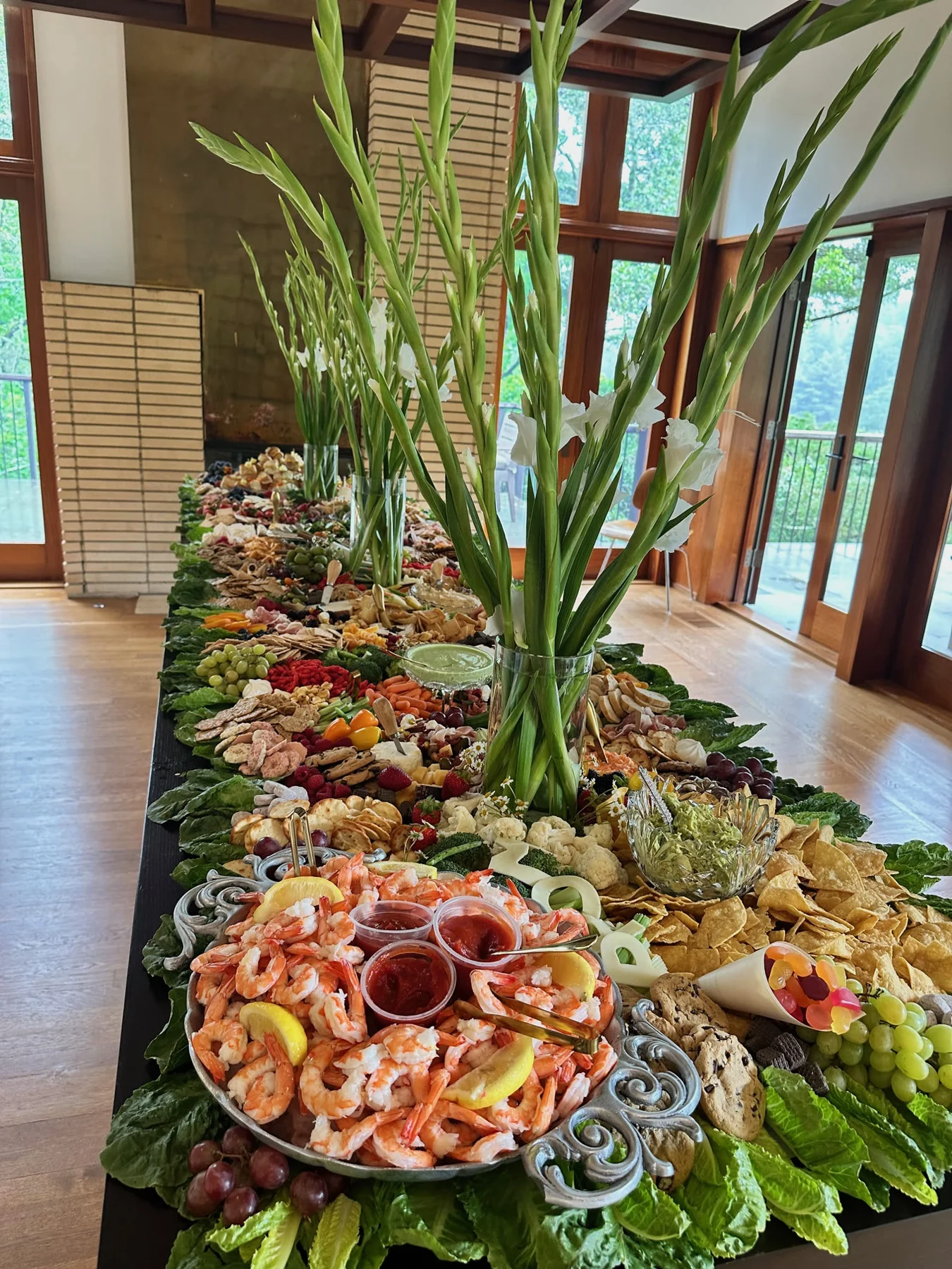 A full-length black prom table with cascading leafy borders, abundant food, a shrimp platter, and tall white gladiolus in clear vases